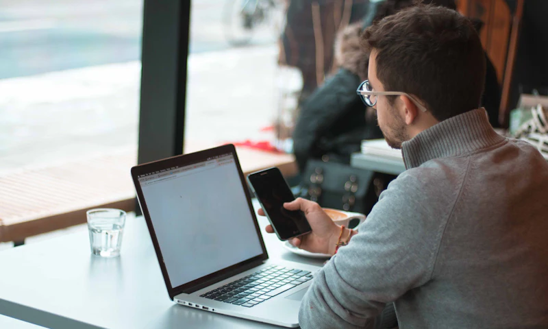 An IT manager working on a laptop and phone by a window